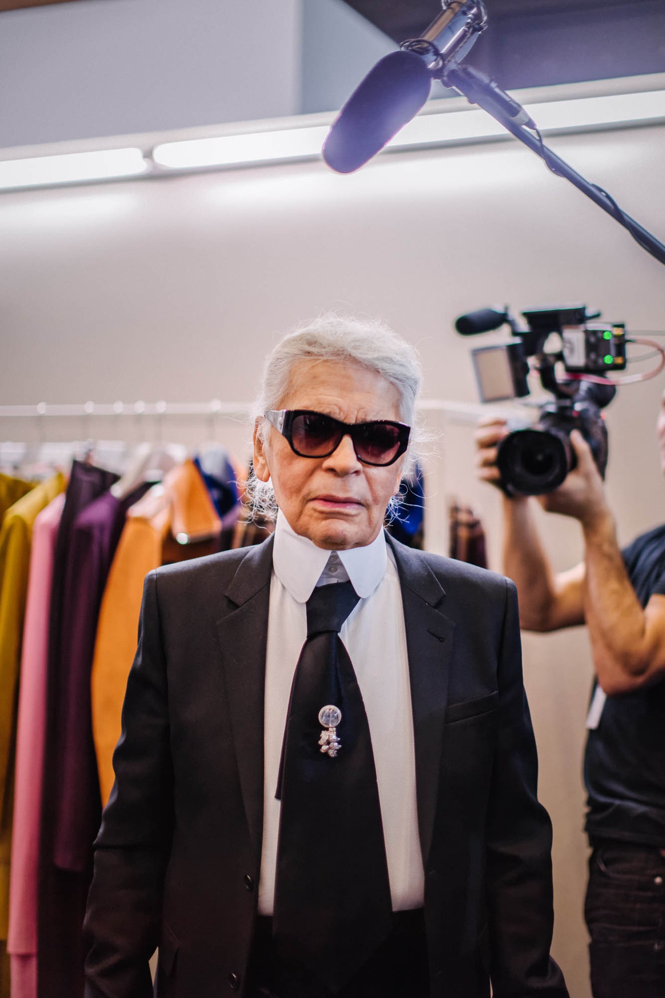 A white-haired man in sunglasses and a dark suit, beside clothing rails and a camera.