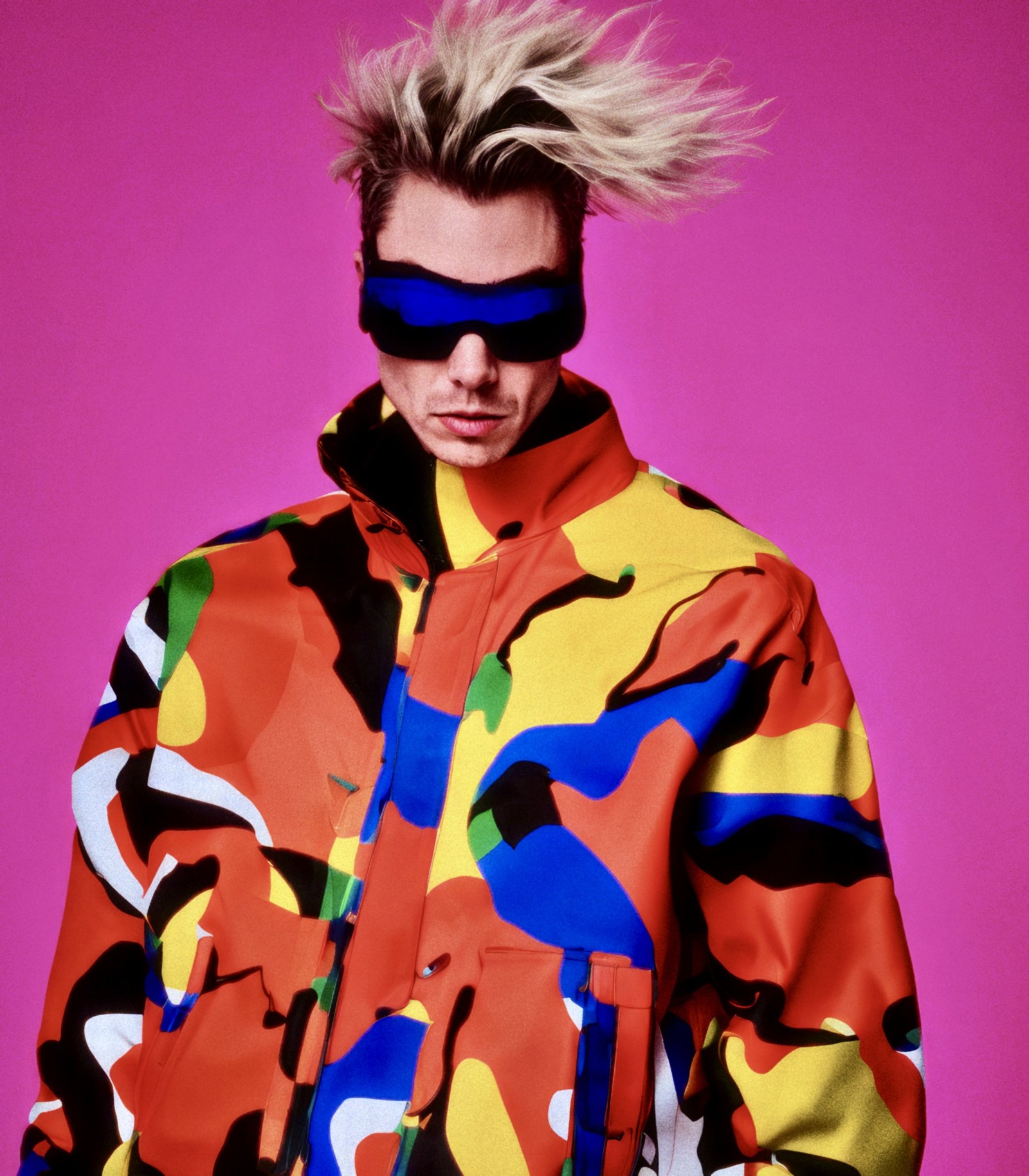 WORM wearing dark glasses and a flowing multicoloured coat against pink.