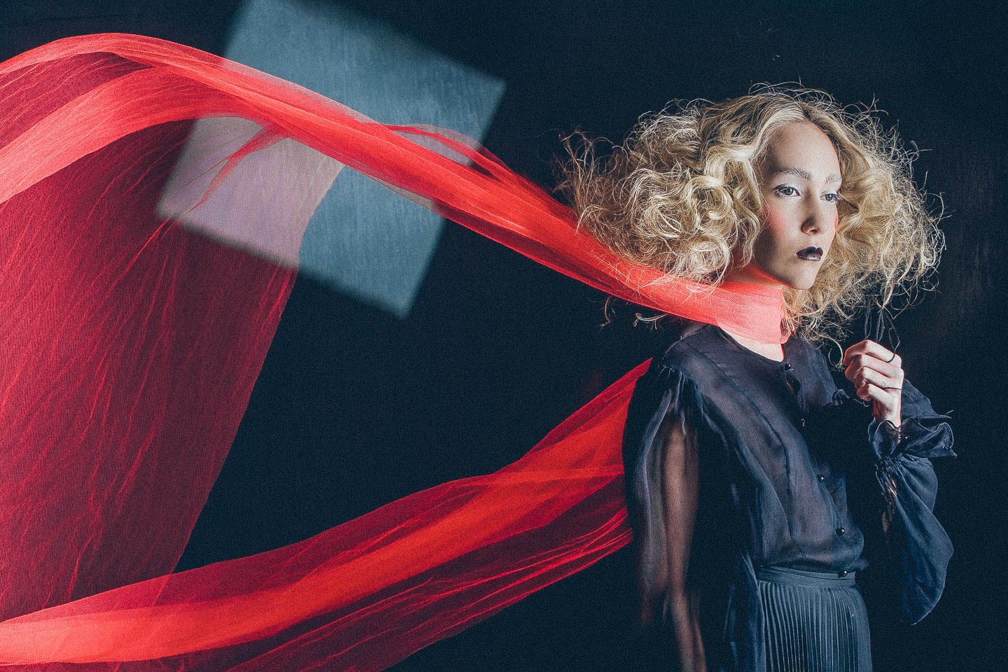 A model in dark clothing with a long red scarf sweeping across the image.