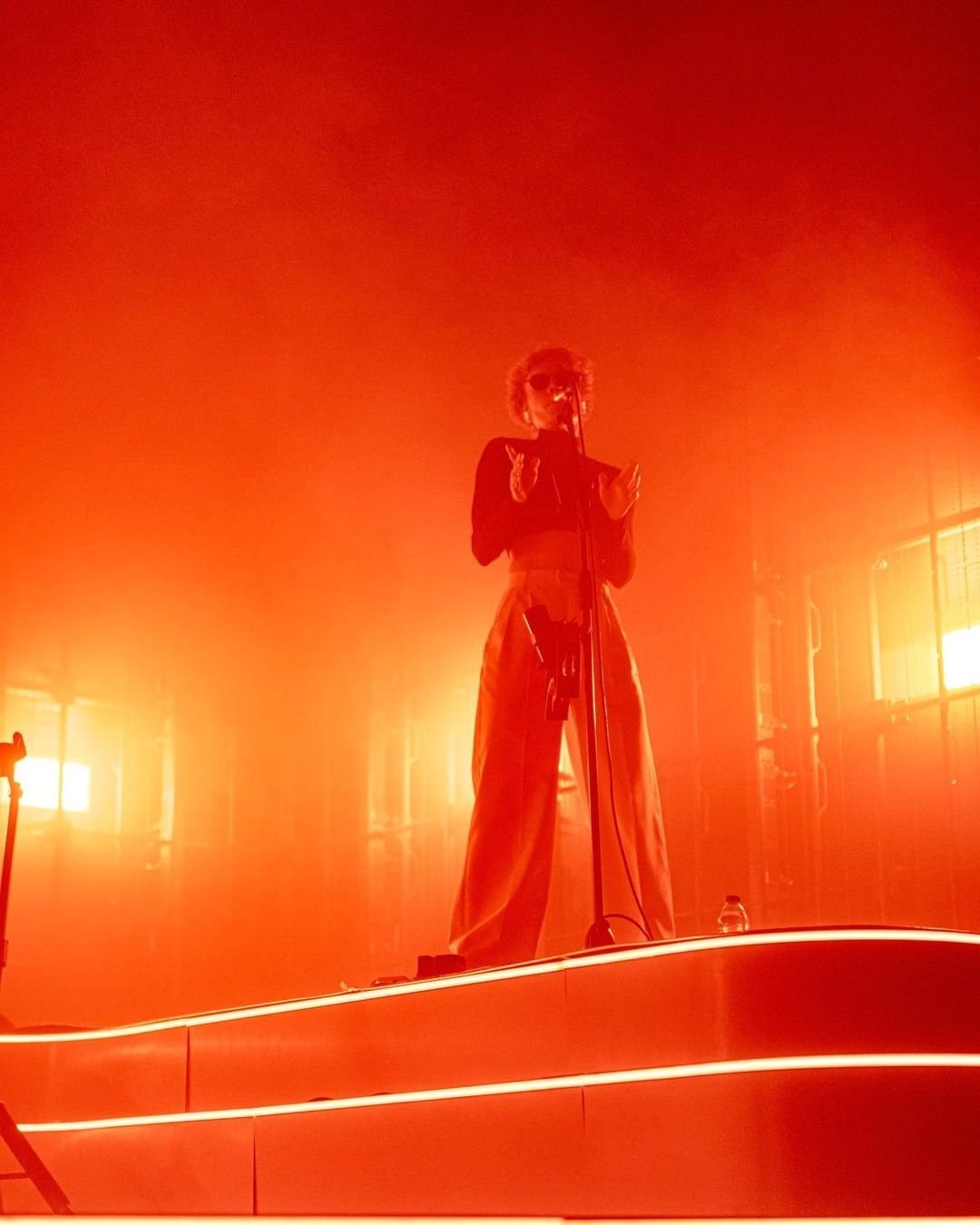 A performer standing at a microphone in orange light and haze.