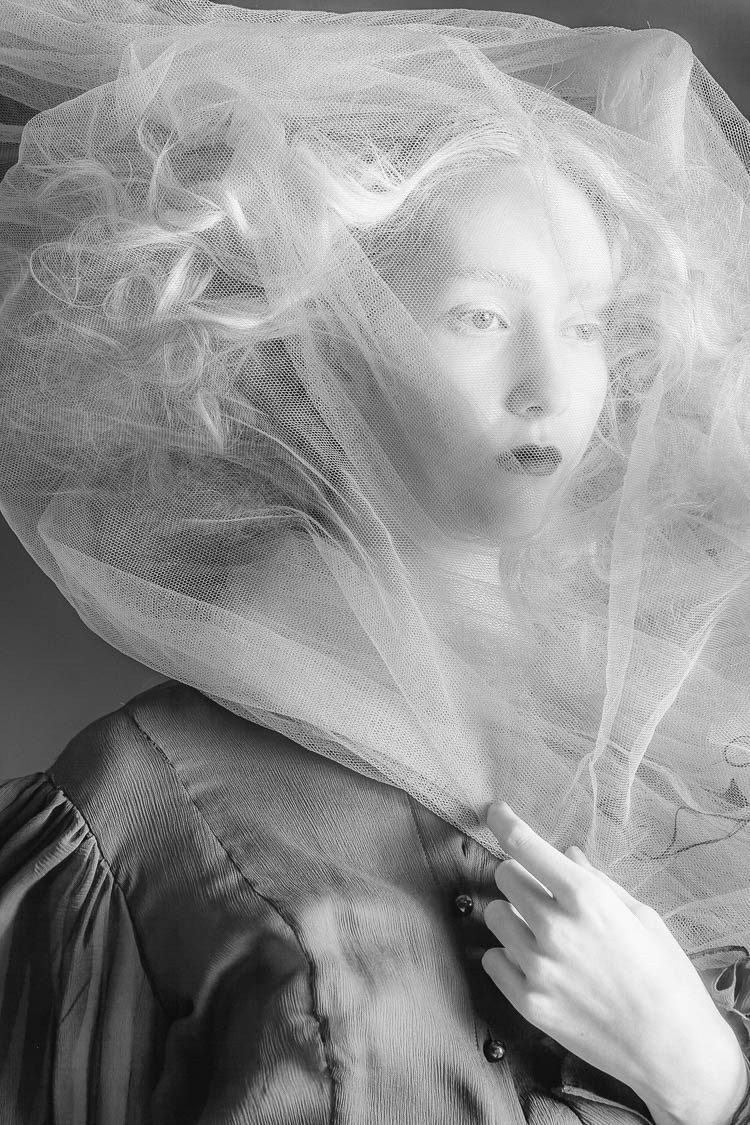 A soft black-and-white portrait of a model behind translucent flowing fabric.