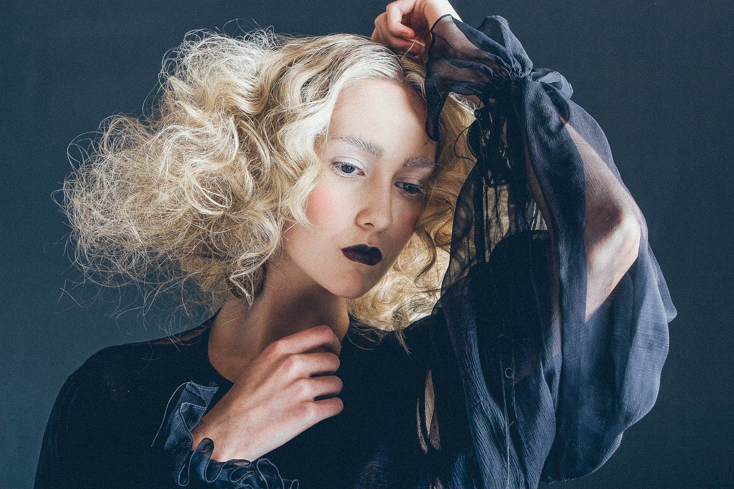 Close portrait of a model with pale curls holding sheer dark fabric above their head.
