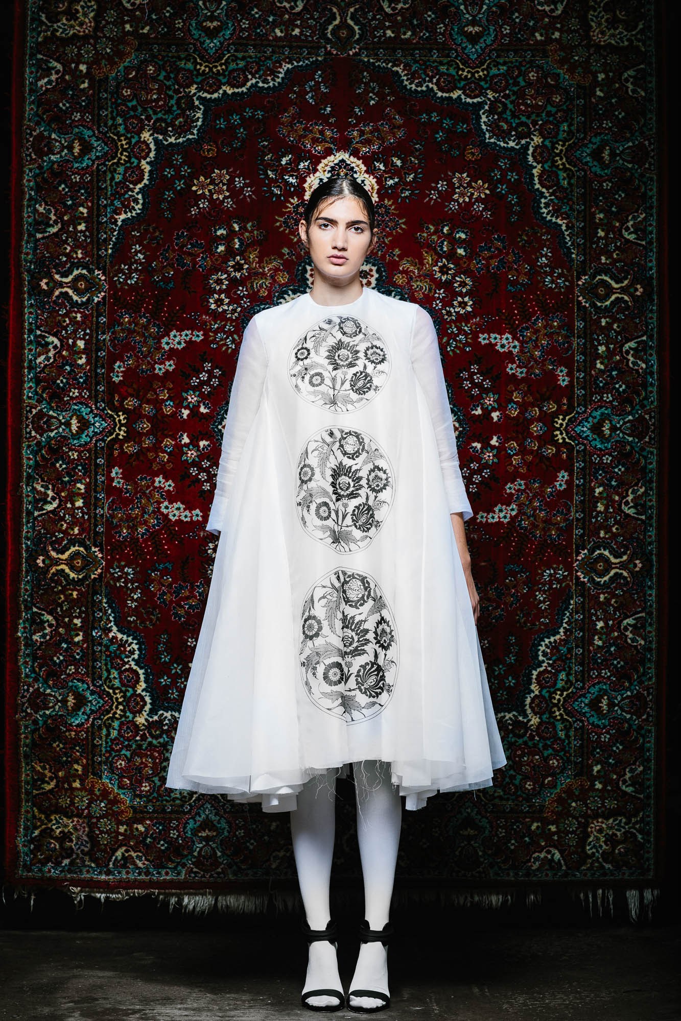 A model in a pale printed dress standing before an ornate red and black textile.
