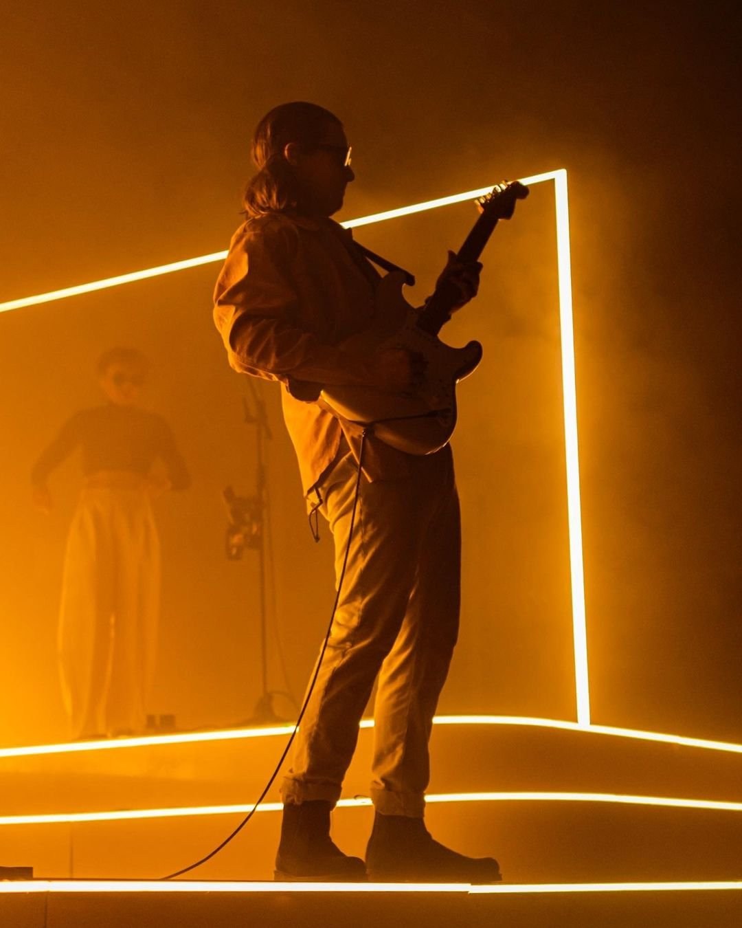 A standing musician holding a guitar under golden stage lighting.