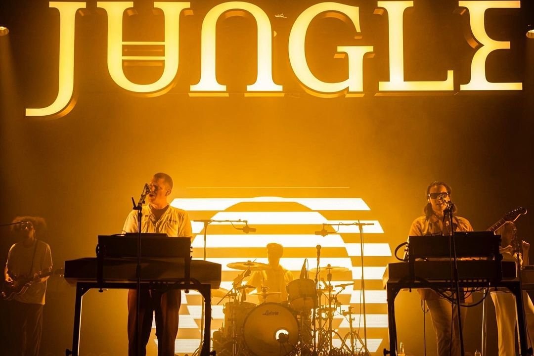 Musicians at keyboards on a stage lit by rows of warm golden lights and JUNGLE lettering.