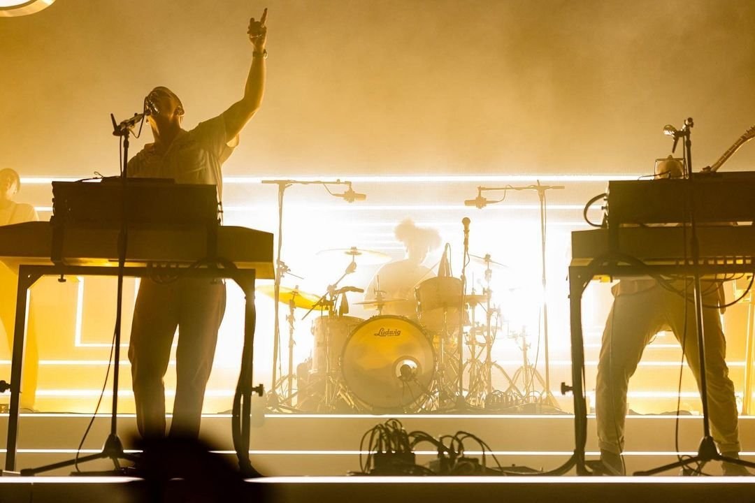 A performer behind a keyboard framed by bright golden stage lights.