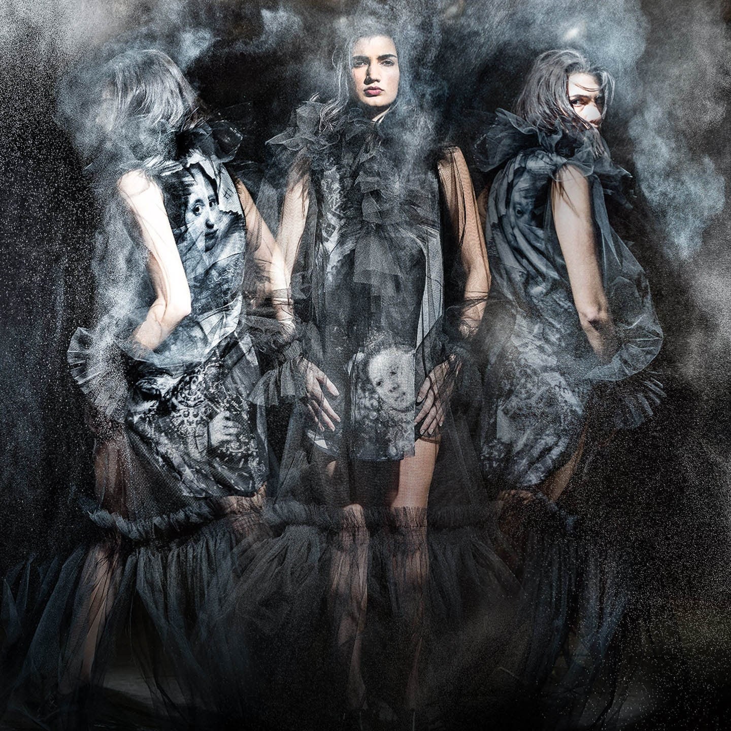 Three models in dark sculptural dresses surrounded by drifting smoke.