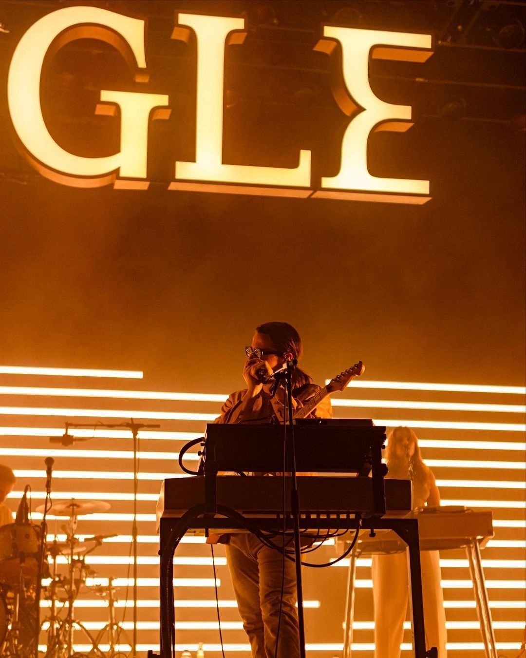 A performer seated at a keyboard against rows of golden lights and oversized lettering.