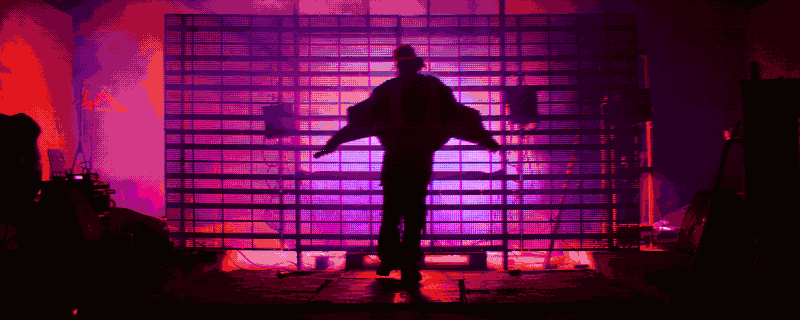 A performer standing with arms extended on a set lit in vivid pink and red.