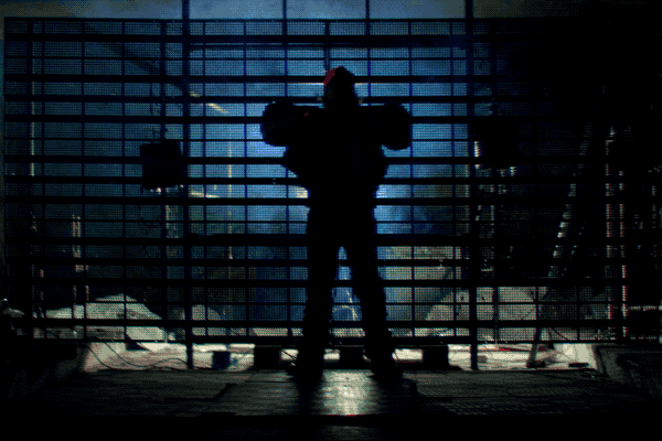 A performer spreading both arms before a metal grid in cool blue light.