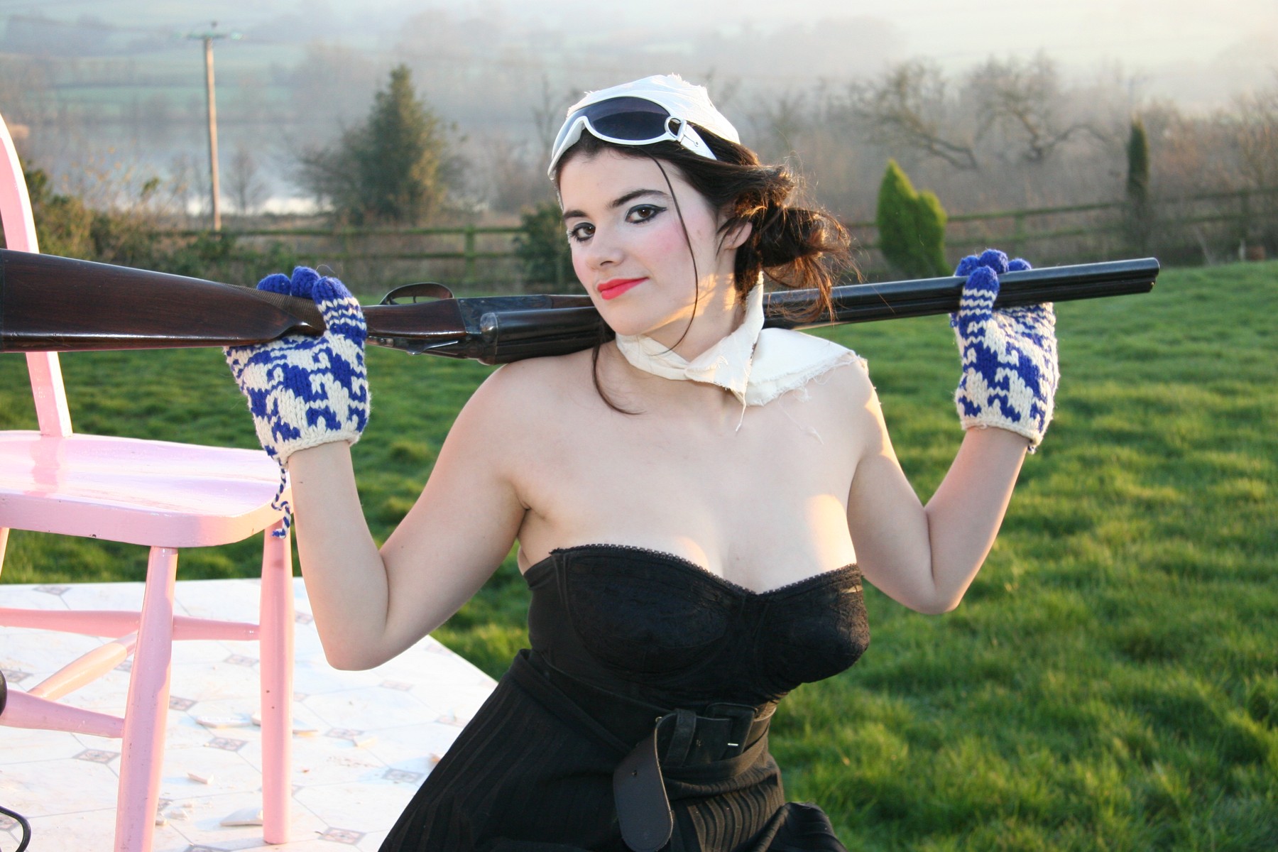 Aviator goggles, patterned gloves and a gun held across the shoulders in a garden portrait.