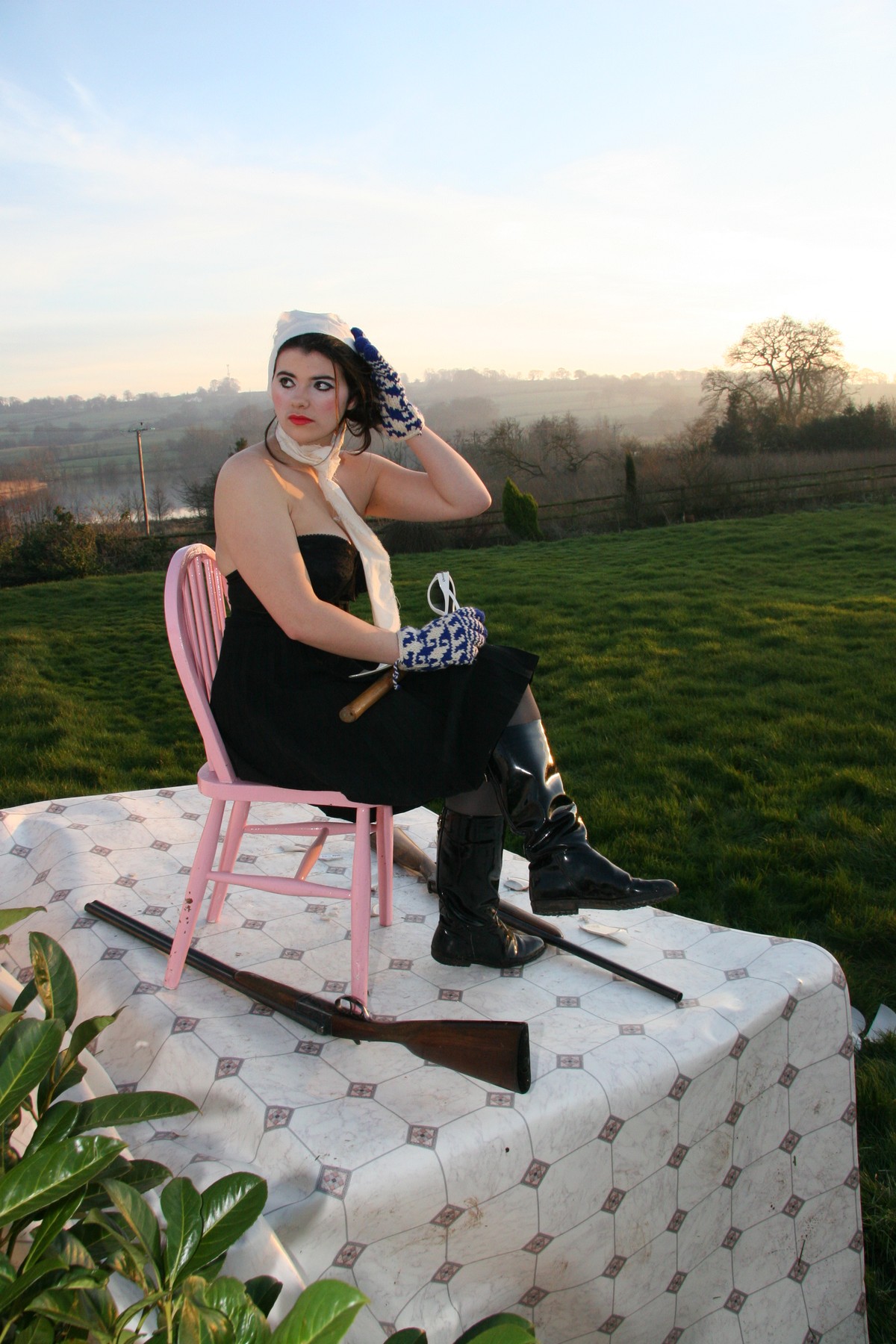 A woman with aviator goggles seated sideways on a pink chair, one hand at her hair.