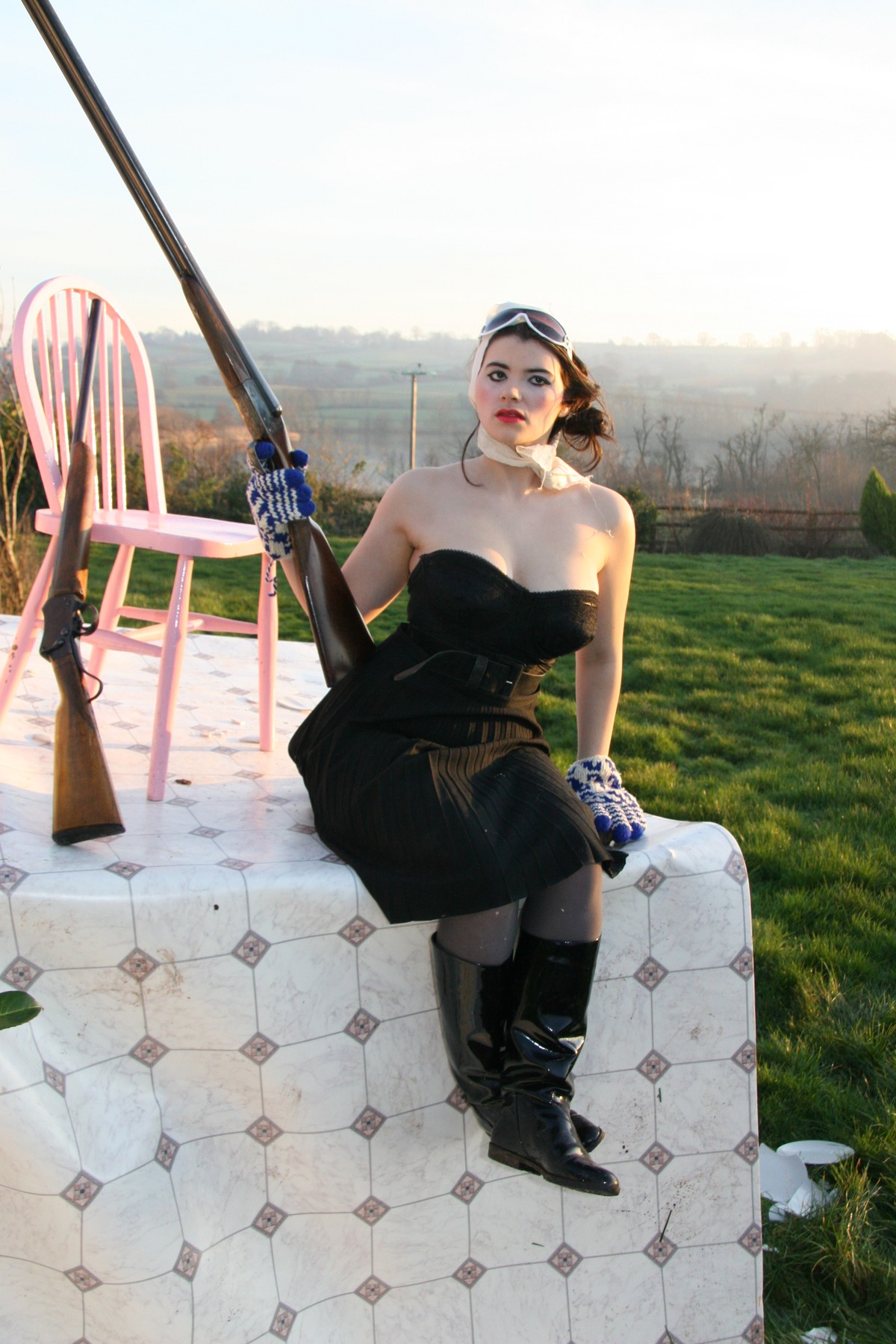 A woman in a black dress and boots seated at the edge of the patterned platform, beside a pink chair.