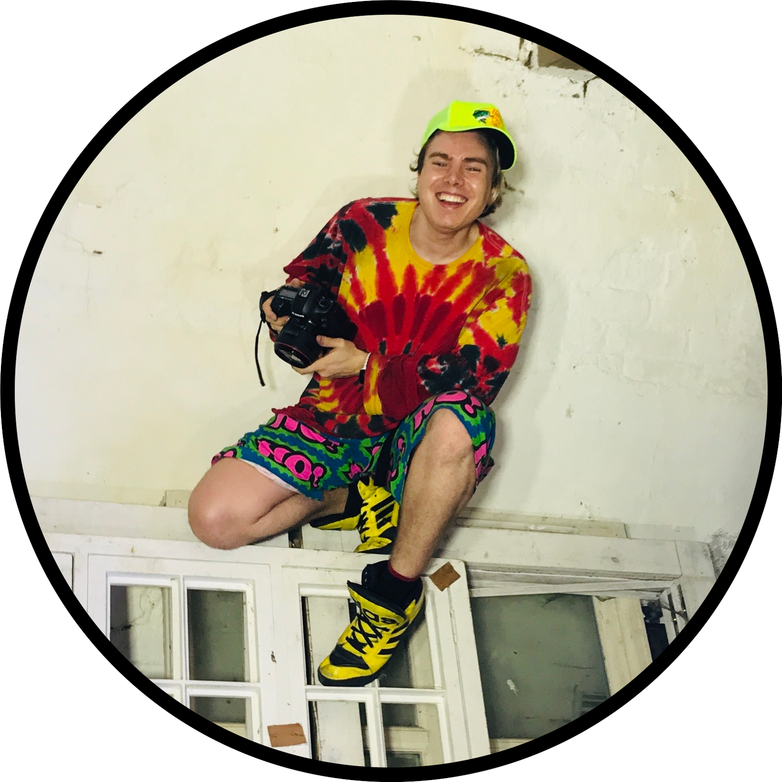 A performer in a multicoloured outfit seated within a circular pale studio backdrop.