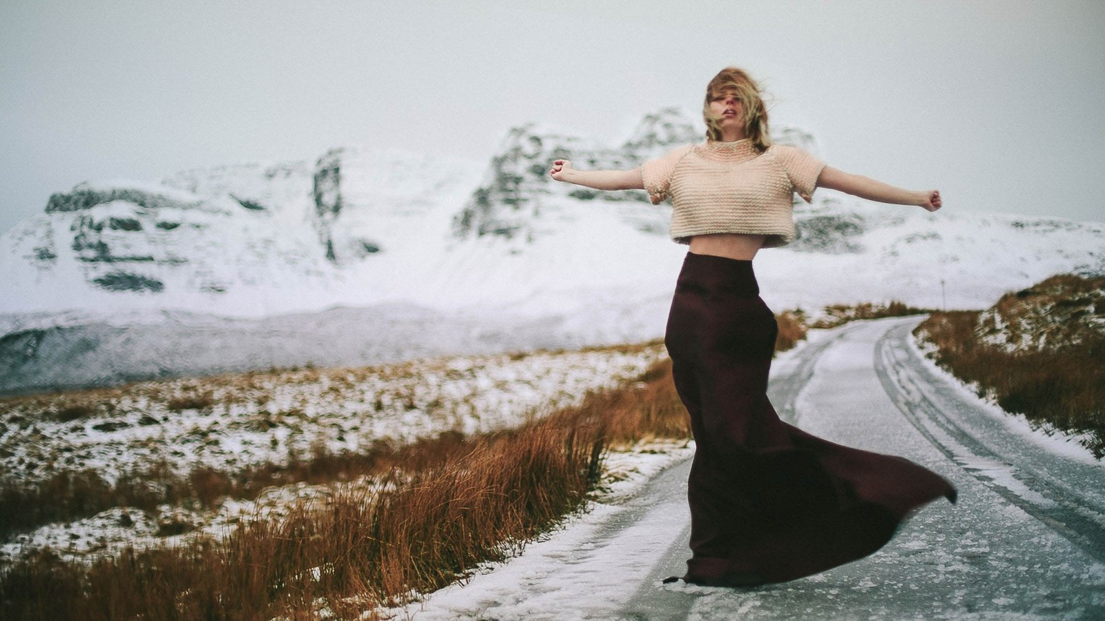 A dancer extending both arms on a winding road through a pale rocky landscape.