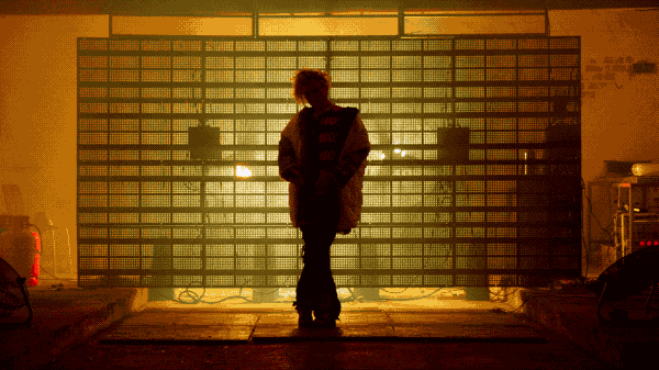 A performer silhouetted against a metal grid illuminated with warm amber light.