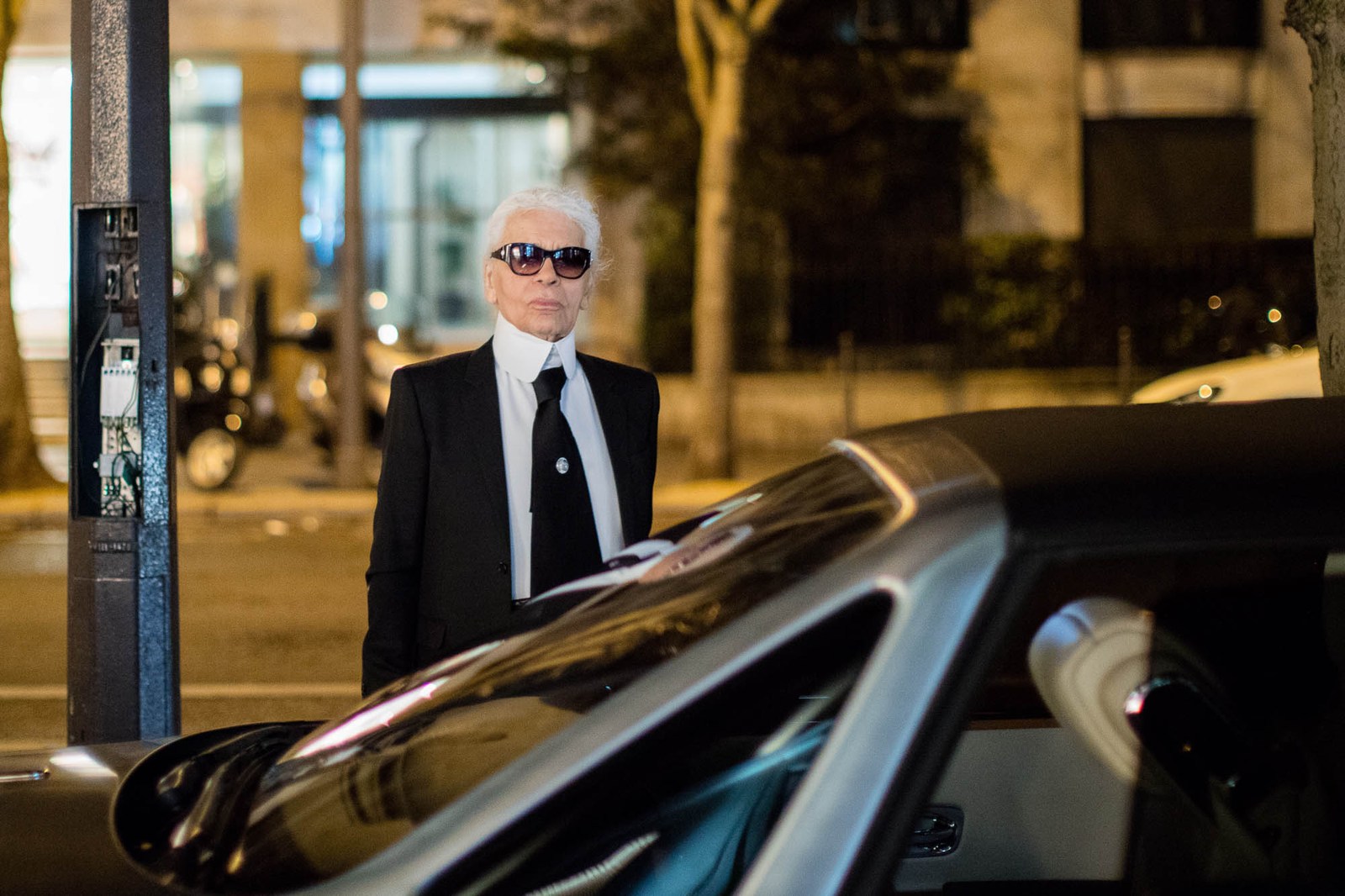 A white-haired man in sunglasses and a suit standing beside a car.