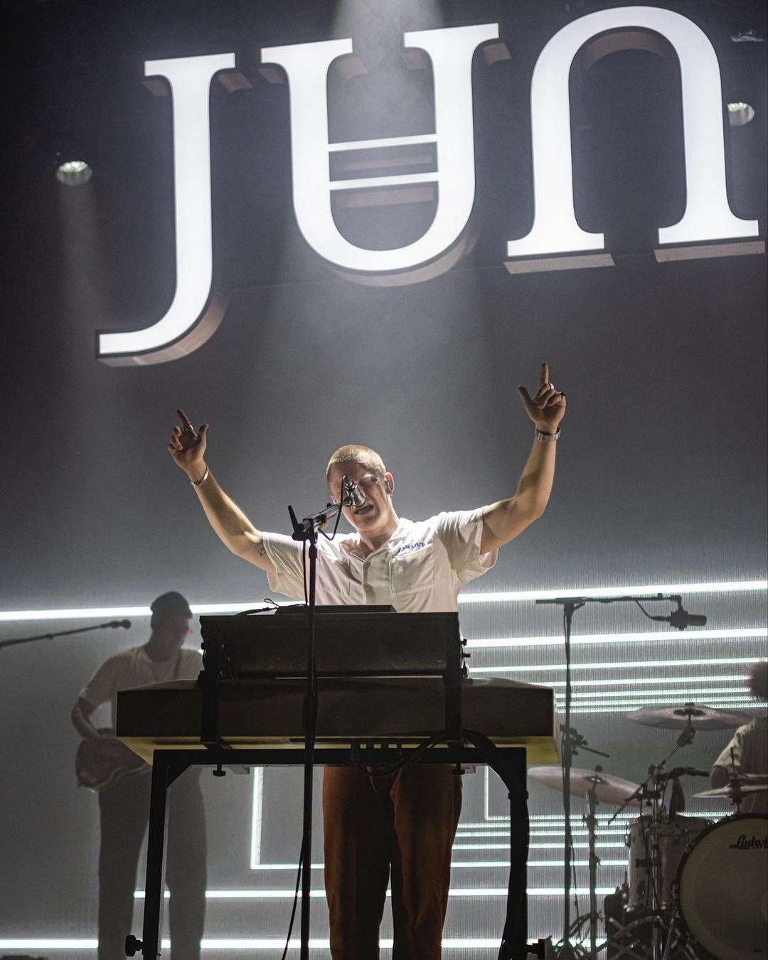 A performer raising both arms behind a keyboard in white stage light.
