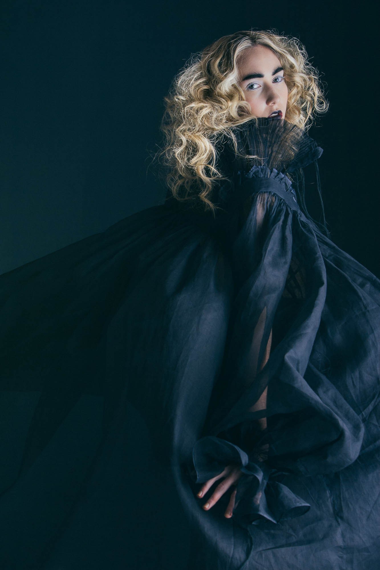 A model with pale curls wrapped in voluminous dark fabric.