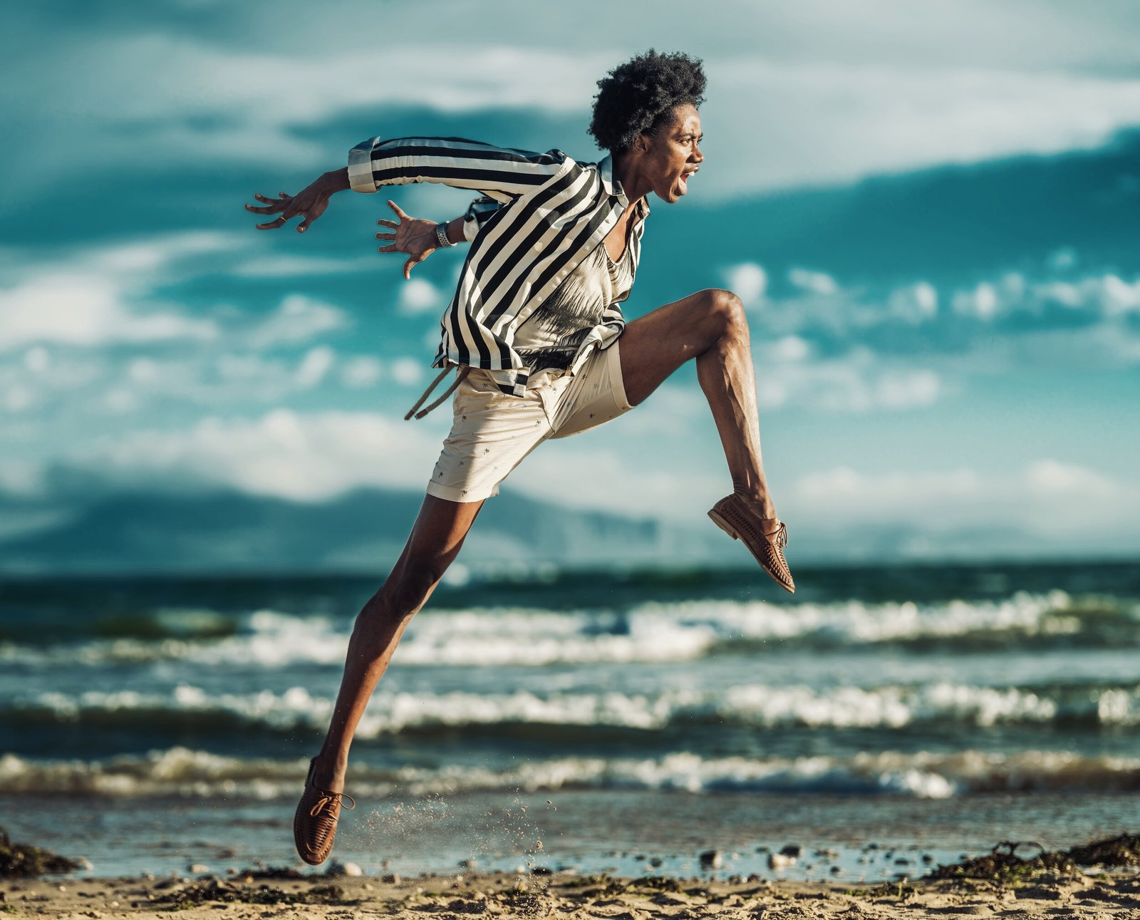 A model in a striped jacket and pale outfit leaping along the shoreline.
