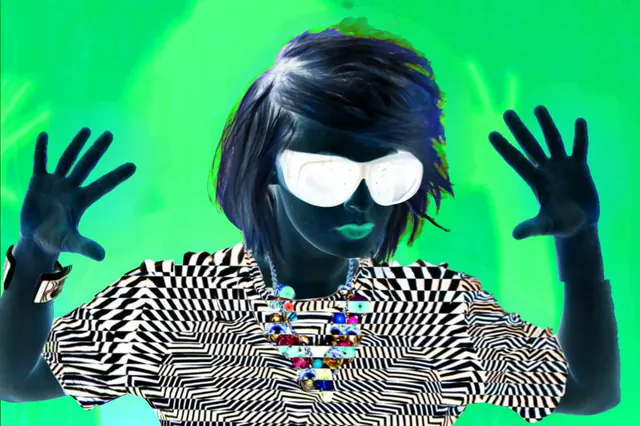 A figure in mirrored sunglasses and a patterned jacket, with both hands raised against vivid green.