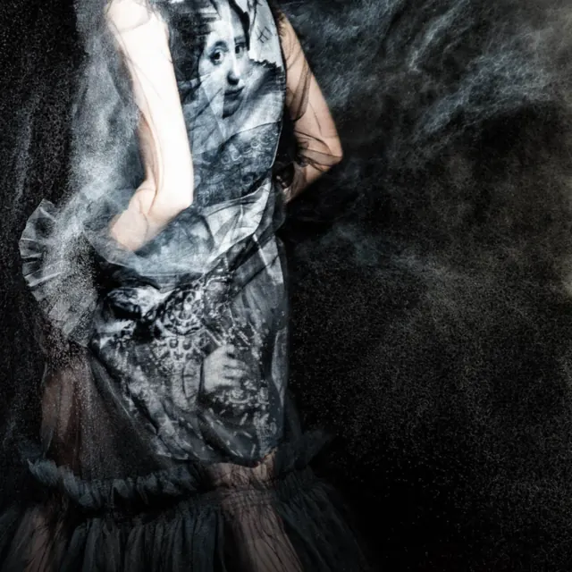 A cropped figure wearing a dress printed with faces amid a cloud of fine grey particles.